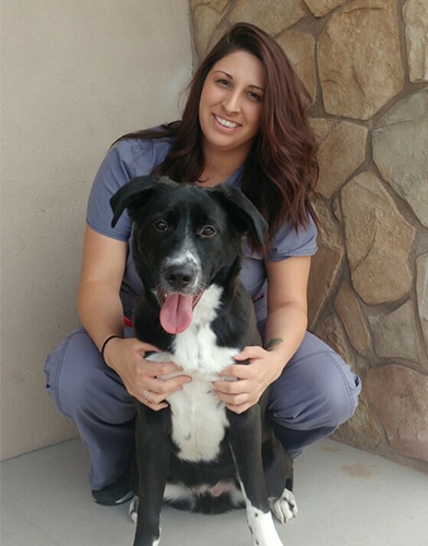 A woman with long brown hair in gray scrubs smiles while sitting on the ground, holding a black and white dog with its tongue out in front of a stone wall.