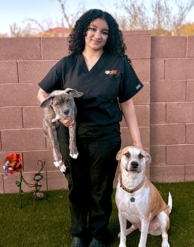 A woman in black scrubs stands outside on grass, holding a gray puppy in one arm and standing next to a large, brown-and-white dog. A brick wall and garden decorations are visible in the background.