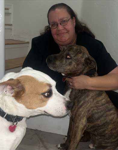 A woman with long hair and glasses sits on stairs, smiling, with her arms around two medium-sized dogs—one brown and one white with brown patches.