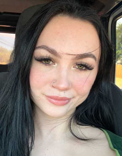 A young woman with long dark hair and light makeup smiles slightly while sitting in a car. Sunlight shines through the window in the background.
